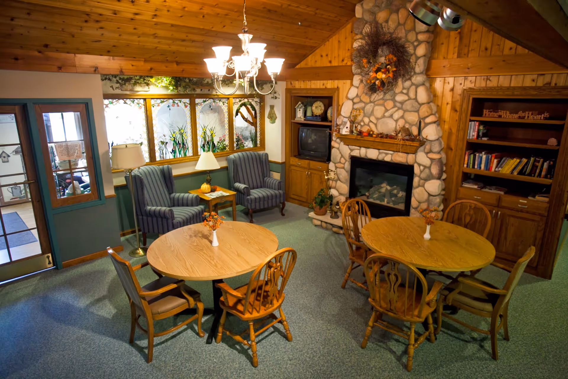 Cozy common area with wooden ceiling and carpeted floor featuring two round wooden tables each surrounded by four chairs. There are two striped armchairs with a small table and lamp between them near a window with decorative stained glass. A stone fireplace with a wreath above it is flanked by wooden built-in shelves, one holding a TV and the other filled with books and decor. A chandelier hangs from the ceiling providing warm lighting.
