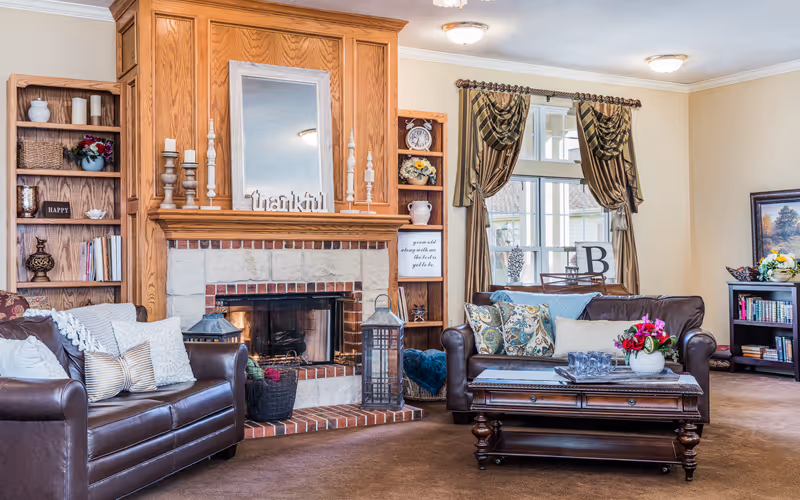 A warm living room with a central fireplace flanked by built-in shelves, leather sofas, a coffee table, and a window with draped curtains.