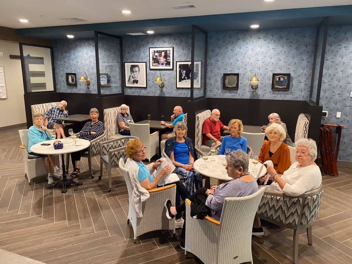 A group of elderly people sitting around two circular tables and several booth-style seats in a common area with patterned blue wallpaper and framed black and white photos on the wall. Some have drinks in front of them and are engaged in conversation.