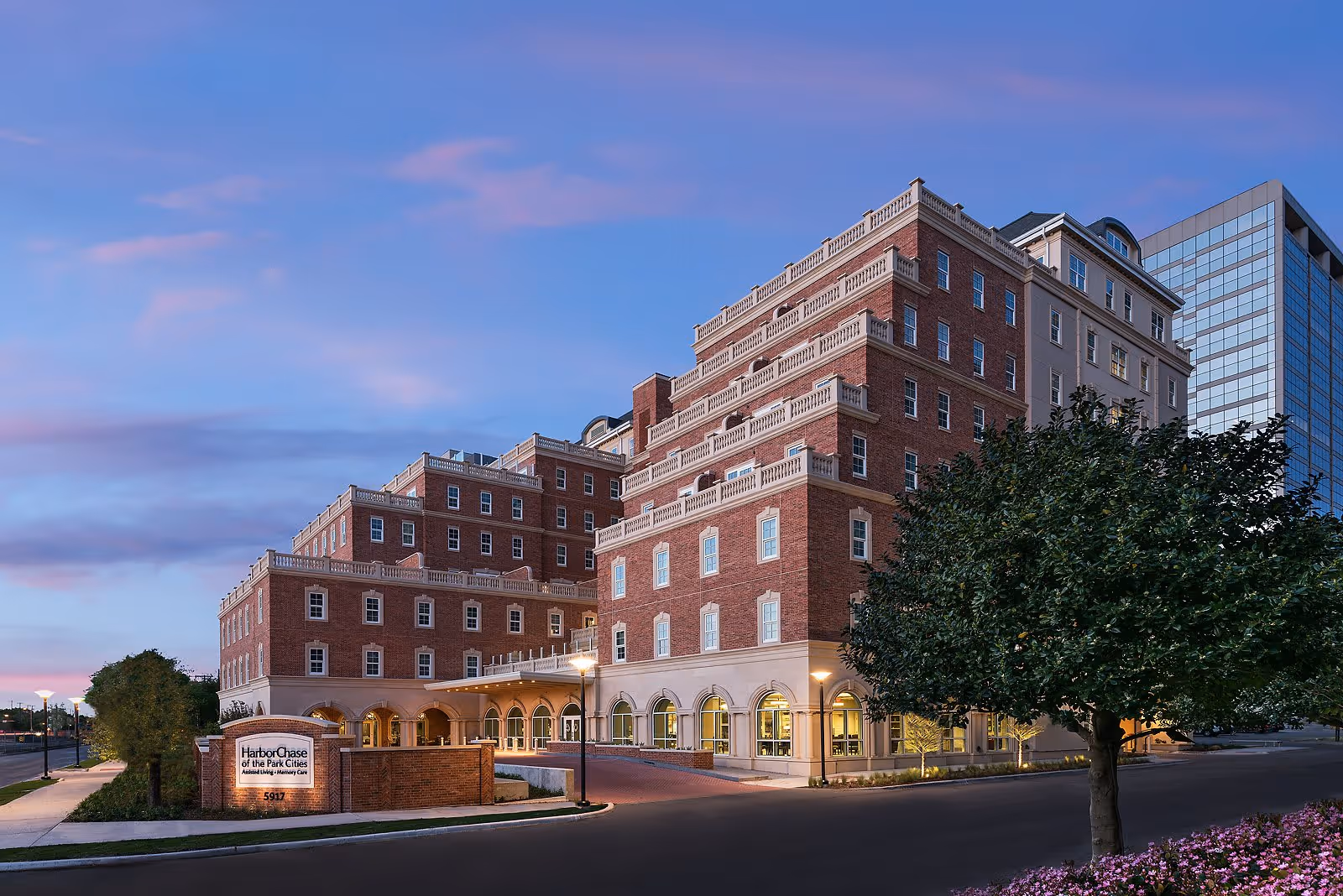 Exterior view of a large multi-story brick building with arched windows and balconies, surrounded by trees and landscaping, under a twilight sky.