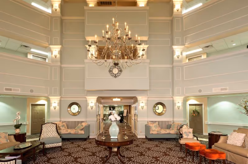 Spacious and elegant common area in a senior living facility with high ceilings, a large chandelier, patterned carpet, multiple seating arrangements including sofas and chairs, decorative mirrors on the walls, and a central round table with a vase of flowers.