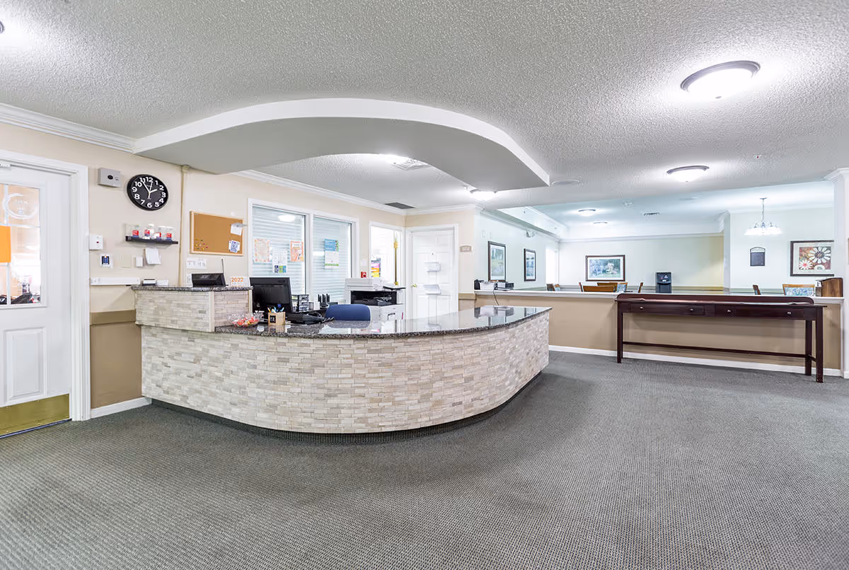 Reception area of a senior living facility with a curved stone front desk, office chair, computer monitors, and a clock on the wall showing 11:50. The room has beige walls, carpeted floor, and ceiling lights. There are framed pictures on the far wall and a long wooden table against a half wall.