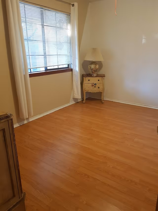 Empty room with wood floors, a window with blinds and curtains, and a small painted nightstand holding a lamp in the corner.