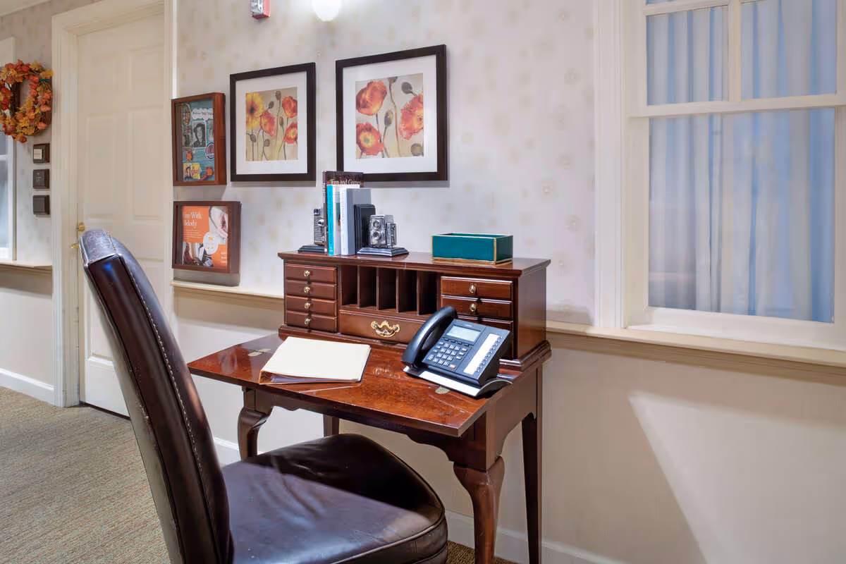 A small wooden desk with multiple drawers and compartments sits against a wall with light patterned wallpaper. On the desk are a telephone, some books, and a closed notebook. A dark leather chair is positioned in front of the desk. Above the desk are two framed floral paintings. To the left, there are additional framed pictures on the wall and a closed door. A window with white curtains is on the right side of the desk.
