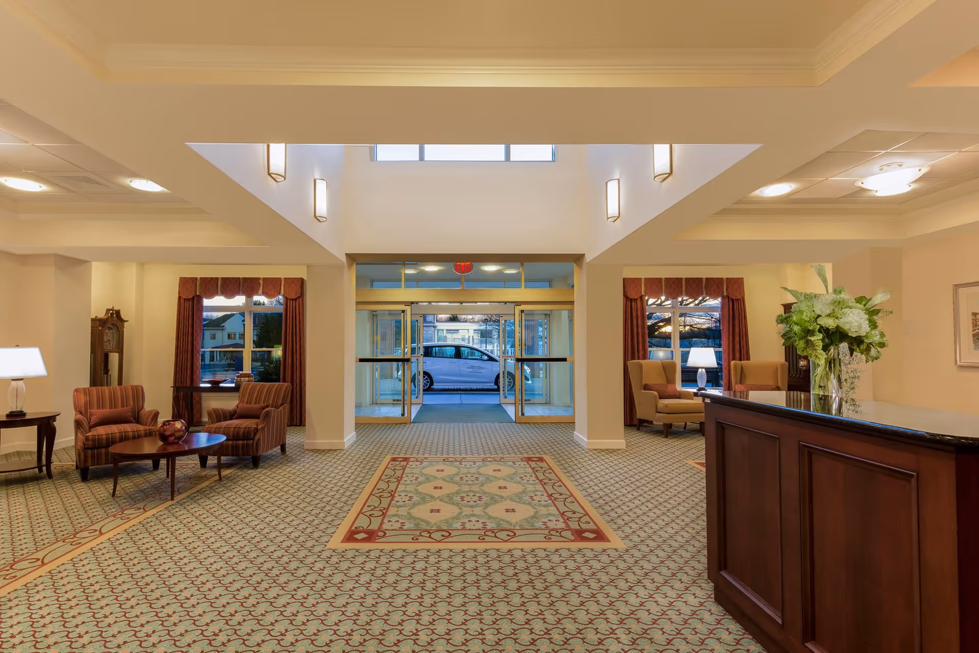 A spacious and well-lit lobby area with patterned carpet, two seating areas each with two armchairs and a small table, a wooden reception desk with a vase of flowers, large windows with red curtains, and glass double doors leading outside where a car is visible.