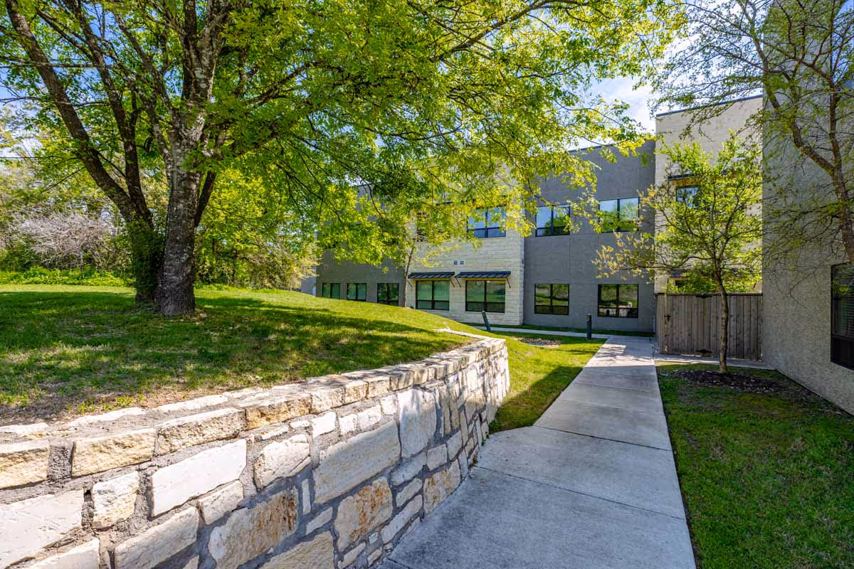A paved walkway beside a stone retaining wall leads to a modern two-story building with large windows. The area is surrounded by green grass, trees with fresh green leaves, and a wooden fence. The sky is clear and blue, indicating a sunny day.