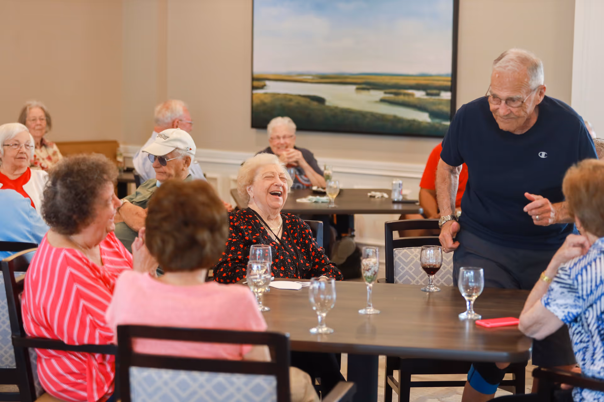 A group of elderly people sitting and standing around tables in a communal dining or activity room, engaging in conversation and laughter. The room has neutral-colored walls and a large landscape painting on the wall.