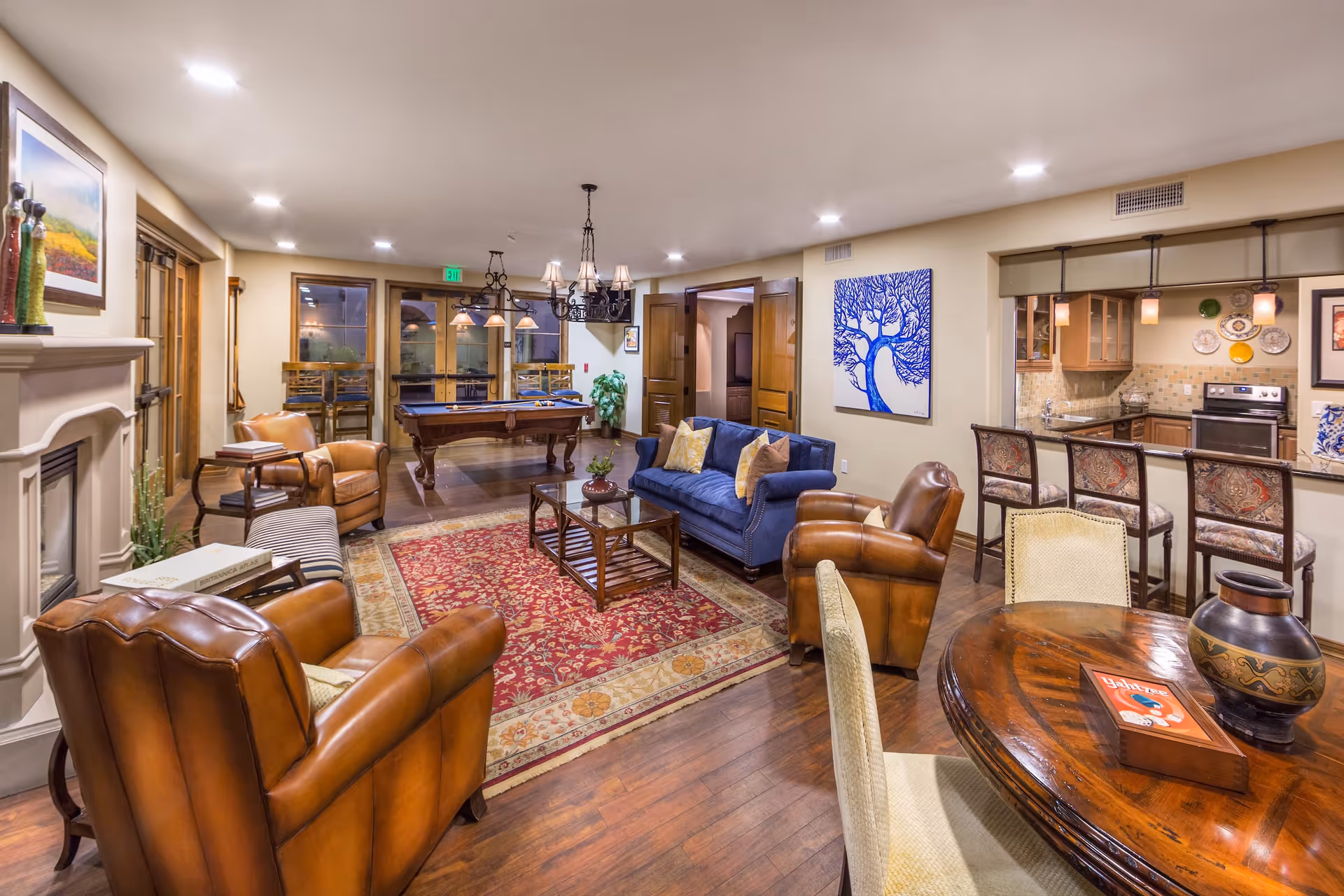 A spacious and warmly lit living area featuring a blue sofa with pillows, several brown leather armchairs, a wooden coffee table on a red patterned rug, and a fireplace on the left. In the background, there is a pool table under hanging lights, a kitchen area with bar stools, and a round wooden table with chairs in the foreground. The walls are decorated with artwork and plates, and the floor is wooden.