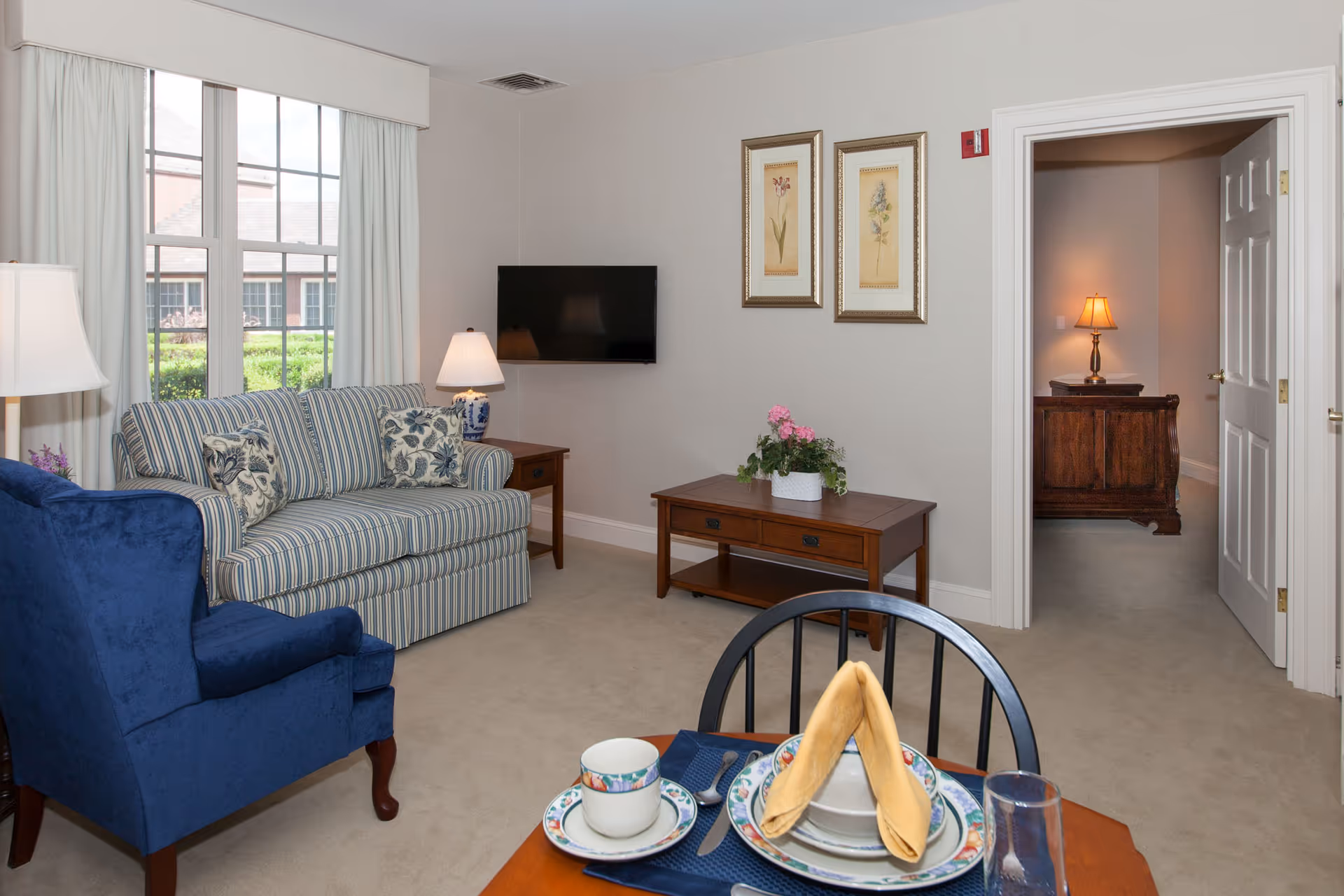 A cozy living room with a striped sofa, blue armchair, wooden coffee table with a potted plant, and a wall-mounted flat-screen TV. There is a window with white curtains letting in natural light. A dining table with place settings is partially visible in the foreground. The room has beige walls and carpet, and an open doorway leads to another room with a wooden dresser and a lit table lamp.