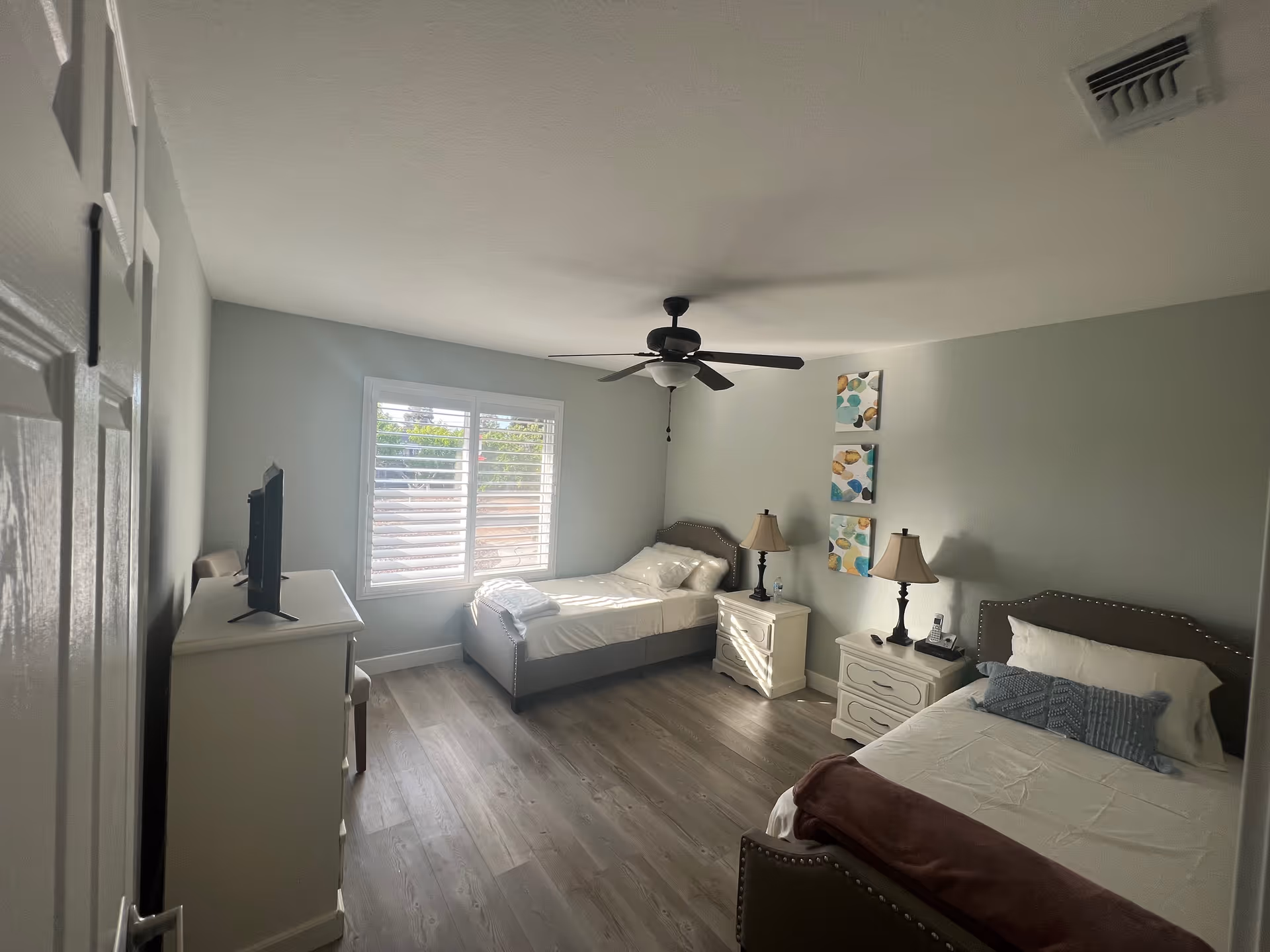 A bright bedroom with two single beds, each with a headboard and bedside table with a lamp. There is a ceiling fan with light in the center of the ceiling. A window with white shutters lets in natural light. A dresser with a TV on top is positioned near the door. The walls are painted light gray and there are three colorful abstract paintings on the wall between the beds. The floor is wood with a light finish.