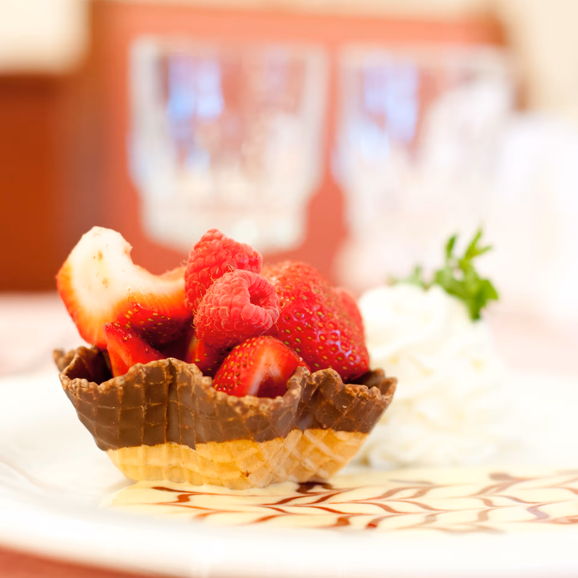 A dessert featuring fresh strawberries and raspberries served in a chocolate-dipped waffle bowl, accompanied by a dollop of whipped cream garnished with a small green herb, presented on a white plate with decorative chocolate drizzle.