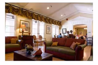 A spacious and well-lit living room area with large windows covered by decorative valances. The room features comfortable seating including a green and red sofa, a wooden coffee table with decorative items, and additional chairs near the windows. The walls are adorned with framed artwork, and there is a lamp on a side table. The room has a high ceiling with recessed lighting and an open archway leading to another area with more seating and decor.