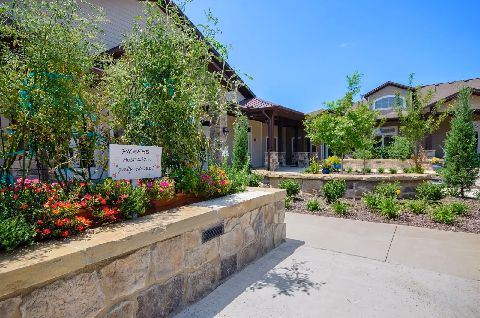 Outdoor garden area at Grand Brook Memory Care of Rogers with stone planters filled with green plants and colorful flowers. A handwritten sign in the garden reads 'PICKERS MUST SAY... pretty please!'. The building with a covered porch and multiple windows is visible in the background under a clear blue sky.