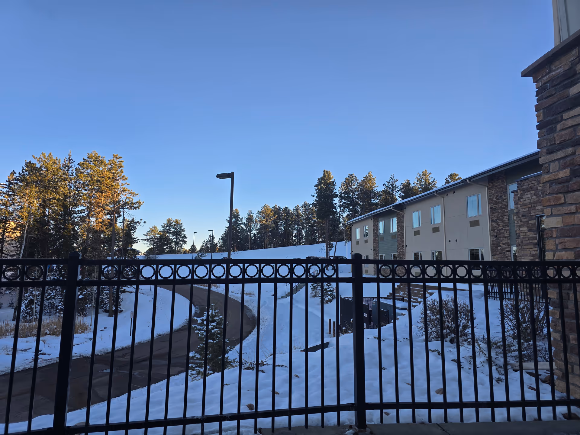 Snow-covered grounds and a curving driveway seen through a black metal fence with trees and a two-story building under a clear blue sky.