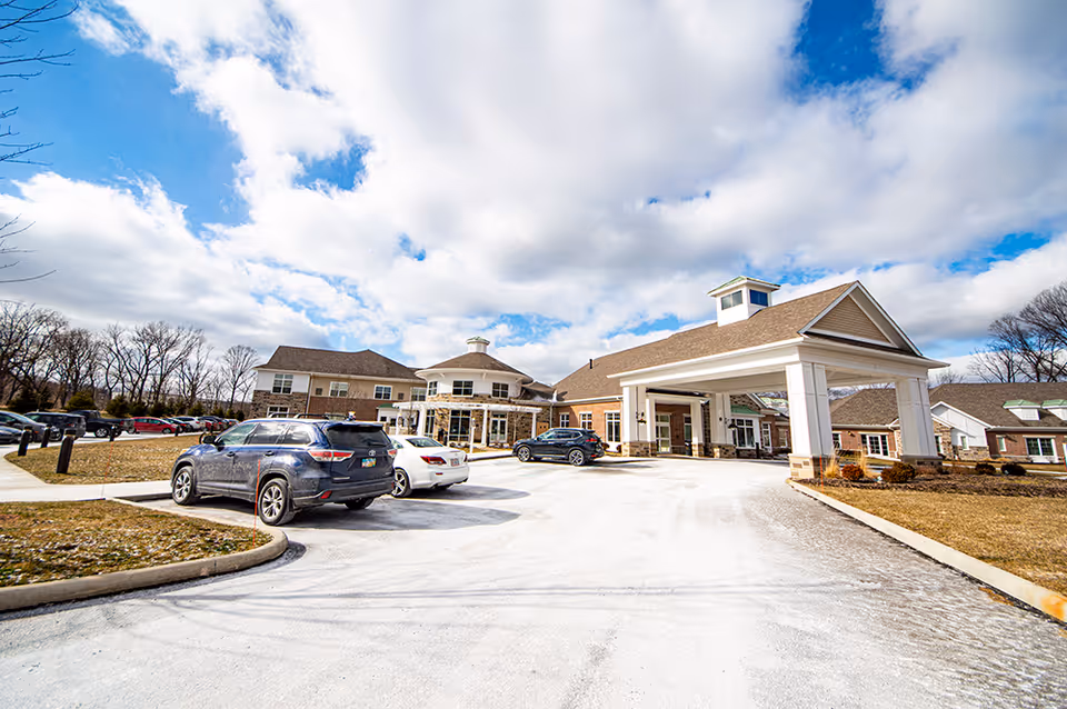 Exterior view of Vista Springs Ravinia Estate senior living facility on a partly cloudy day, showing a large covered entrance with several parked cars and surrounding trees.