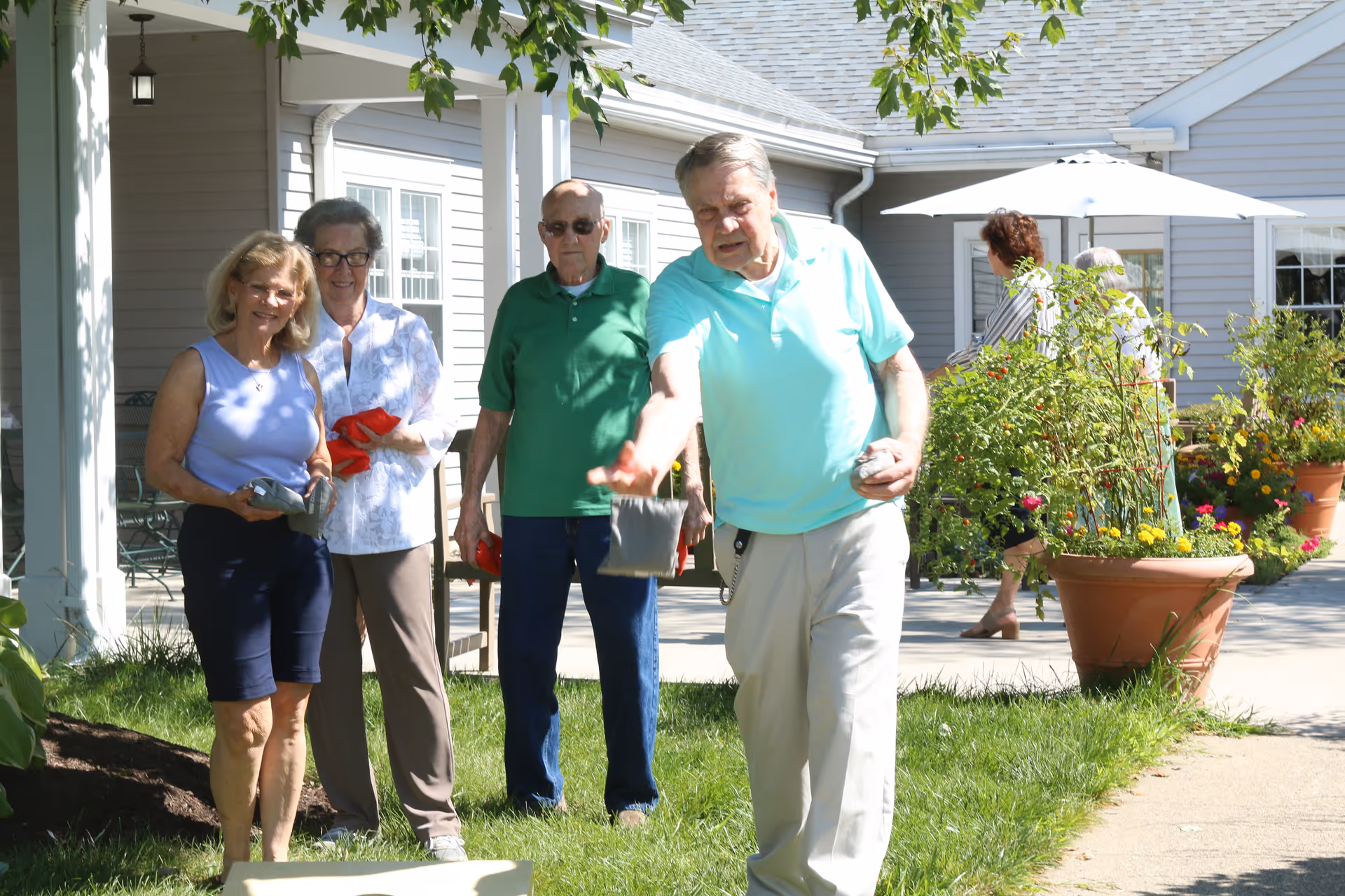 A group of elderly people enjoying an outdoor game of cornhole on the lawn outside a residential building with potted plants and a patio umbrella in the background.