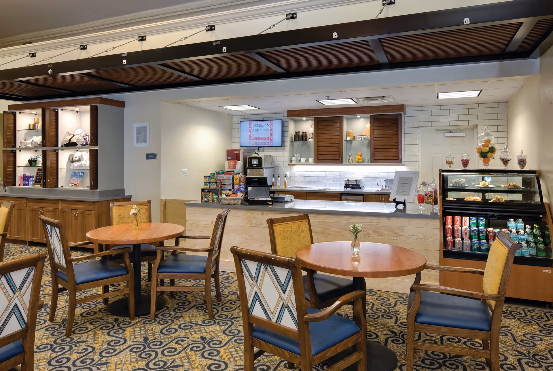 A cozy dining area in a senior living facility featuring round wooden tables with chairs that have patterned backs and blue cushions. The room has a carpet with a decorative pattern. In the background, there is a counter with a display of snacks, a coffee machine, and a refrigerated display case stocked with beverages and pastries. Shelves with decorative items and a small vase with flowers on each table add to the welcoming atmosphere.