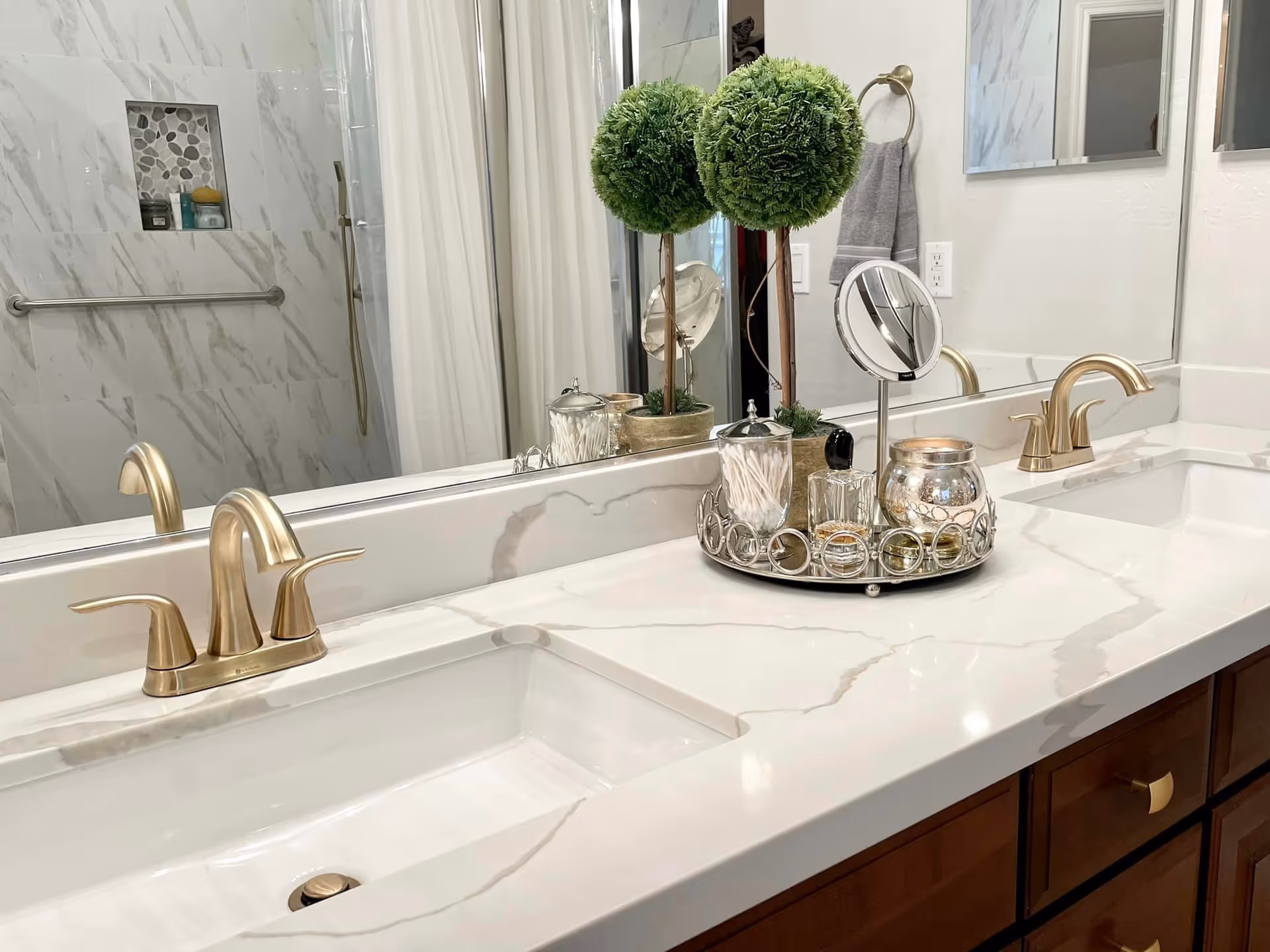 A modern bathroom countertop with a white marble surface featuring two gold faucets and sinks. On the countertop, there is a decorative tray holding a small round mirror, cotton swabs, a candle, and two artificial topiary plants. In the background, a large mirror reflects part of a shower area with a grab bar and a niche containing toiletries.