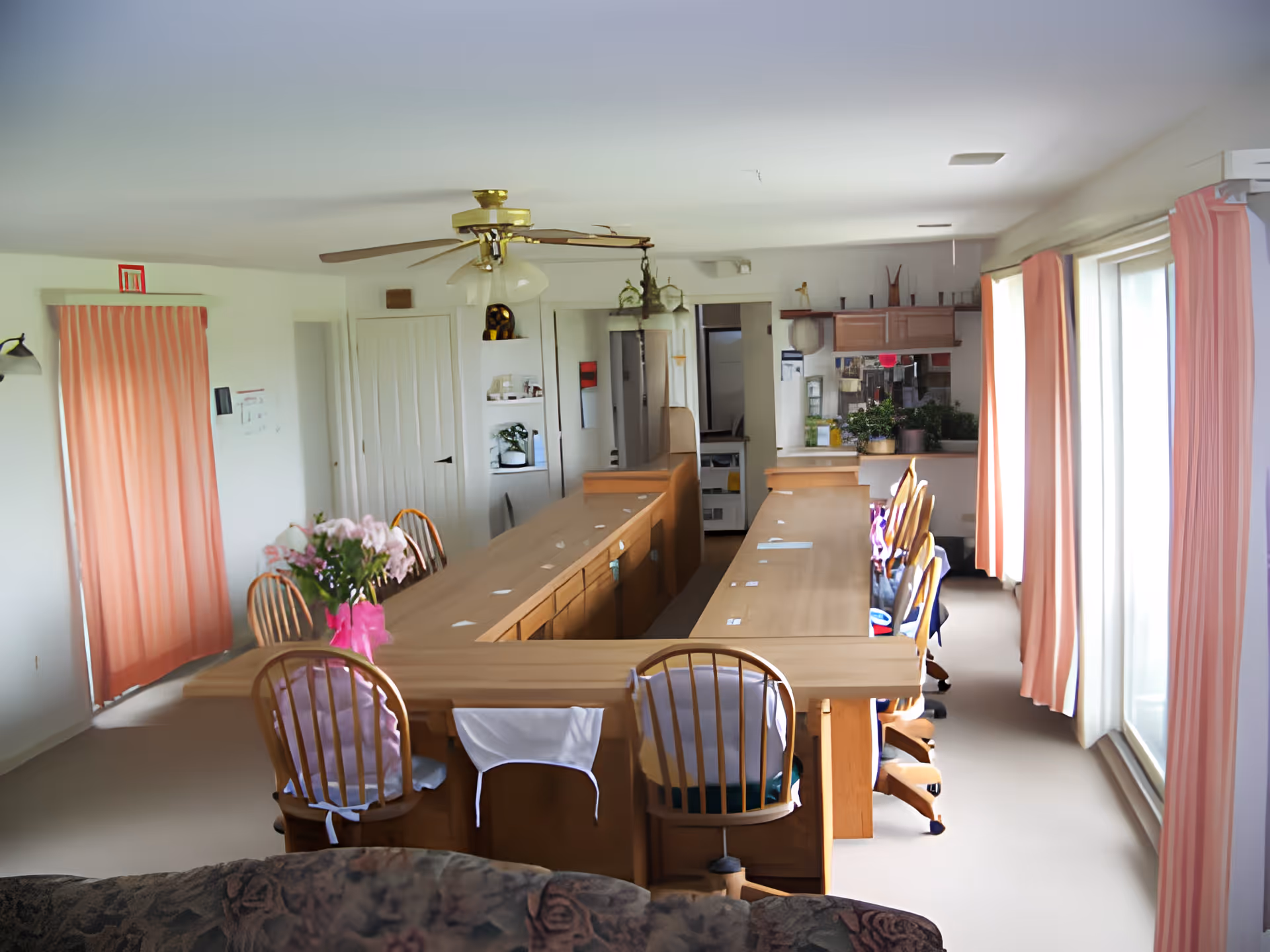 Interior view of a communal dining area with a large U-shaped wooden table surrounded by wooden chairs, some with cushions and cloths draped over the backs. The room has large windows with pink curtains allowing natural light to enter. There is a ceiling fan with lights above the table, and shelves with plants and decorative items in the background. The space appears clean and organized, designed for group dining or activities.