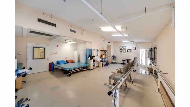 A spacious physical therapy/rehab room with parallel bars, treatment tables, chairs, and medical equipment.