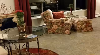 Cozy living room area with a floral patterned armchair and matching ottoman, a glass-top round table with a book on it, a small plant on a stand, and red curtains covering large windows.