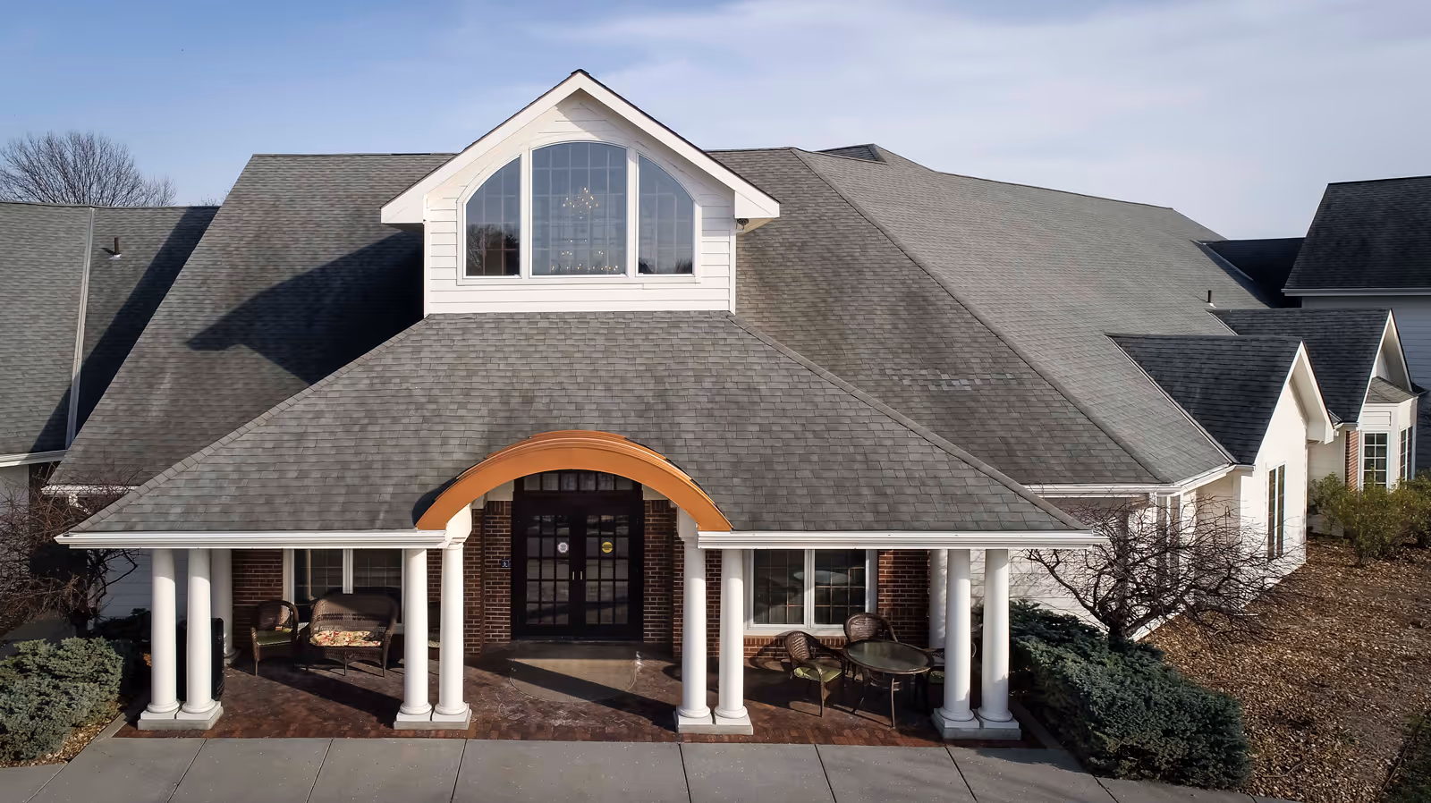 Front exterior view of a senior living facility building with a large gray shingled roof, white pillars supporting a covered entrance with a curved orange awning, and outdoor seating including wicker chairs and a glass table. The building has large windows and is surrounded by landscaping with bushes and trees.