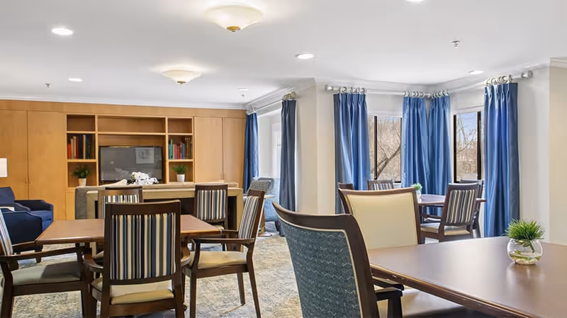 A bright and spacious common area in a senior living community featuring multiple wooden tables and chairs with striped and solid upholstery. Large windows with blue curtains allow natural light to fill the room. A built-in wooden entertainment center with shelves, books, and plants is visible in the background.
