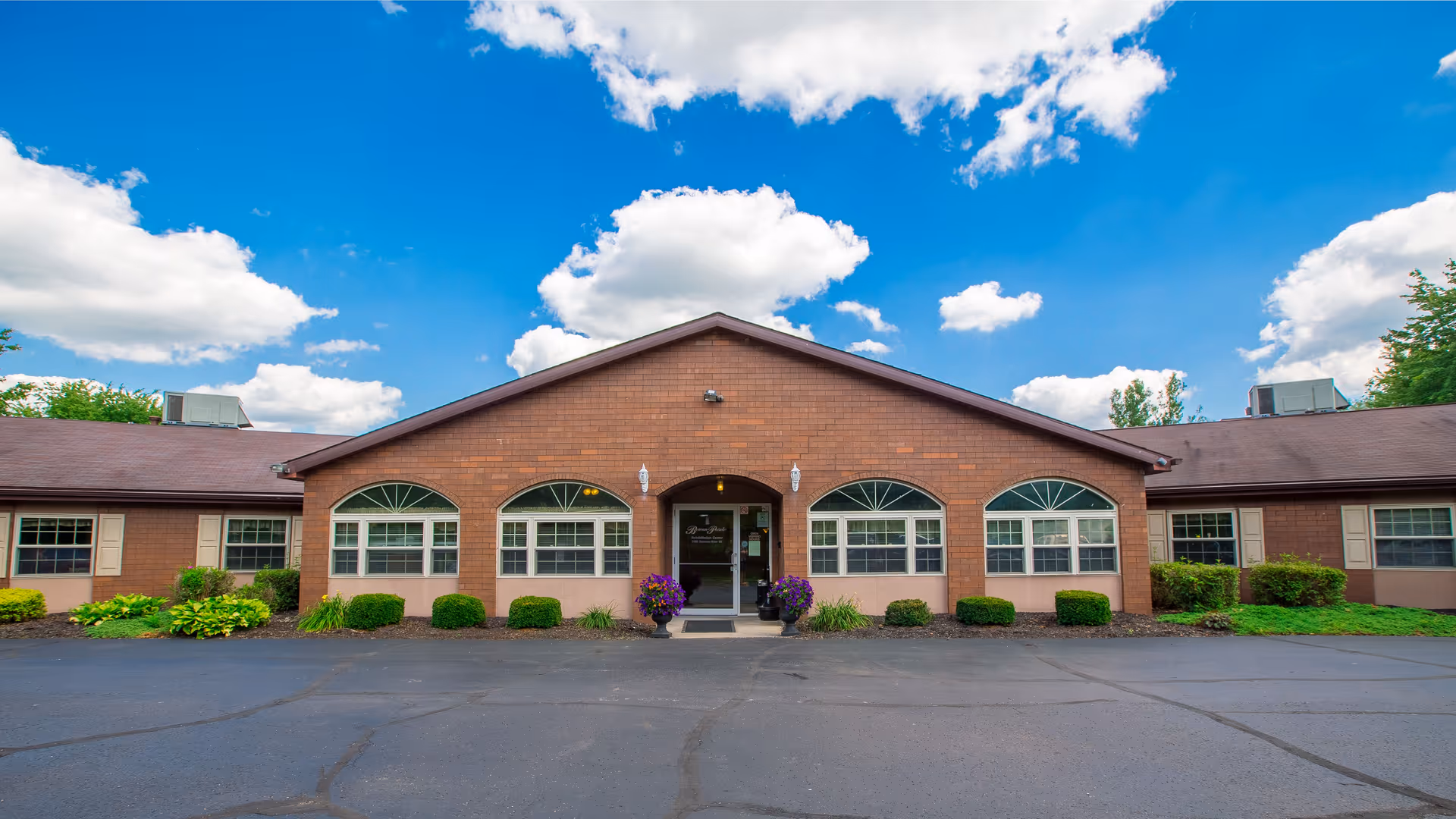 Single-story brick building front with arched windows, a central entrance, shrubs, and a paved driveway under a blue sky with clouds.