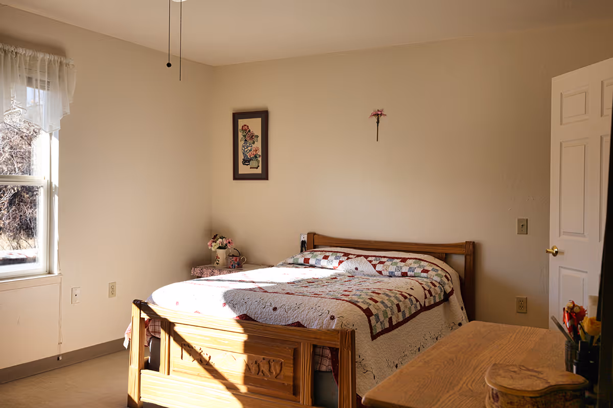 A cozy bedroom with a wooden bed frame and a colorful patchwork quilt. There is a window with sheer curtains letting in natural light, a small bedside table with a vase of flowers, a framed floral picture on the wall, and a wooden dresser in the foreground.