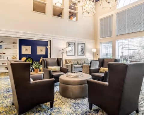 A spacious and well-lit living room area with four dark brown armchairs arranged around a round ottoman. Behind the chairs is a gray sofa with cushions, two framed artworks on the wall, a floor lamp, and large windows with blinds letting in natural light. The room features a high ceiling with hanging light fixtures and a patterned carpet.