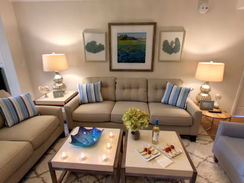 A cozy living room with a beige tufted sofa and matching loveseat, each adorned with striped blue and white pillows. Two side tables with lamps flank the sofa. In front, two square coffee tables hold a blue decorative bowl, a small plant, a bottle of wine, and plates with snacks. Three framed artworks hang on the wall above the sofa.