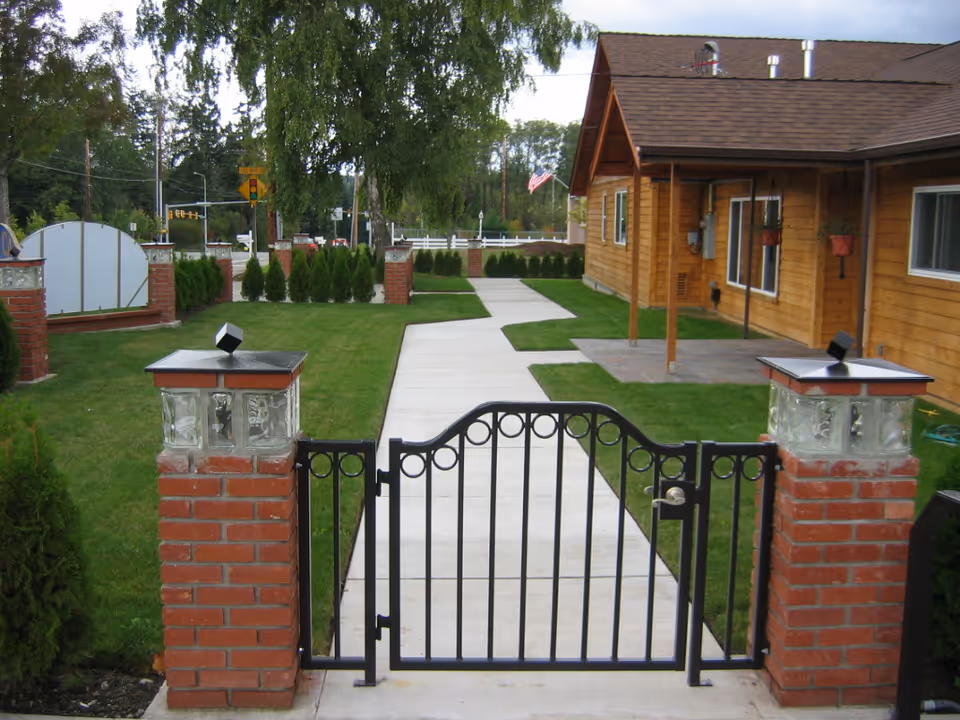 A gated entrance with a black metal gate set between two brick pillars topped with glass block lanterns. A concrete pathway leads from the gate through a well-maintained green lawn towards a wooden building with a brown roof and hanging flower pots. Trees and a street with traffic lights are visible in the background.