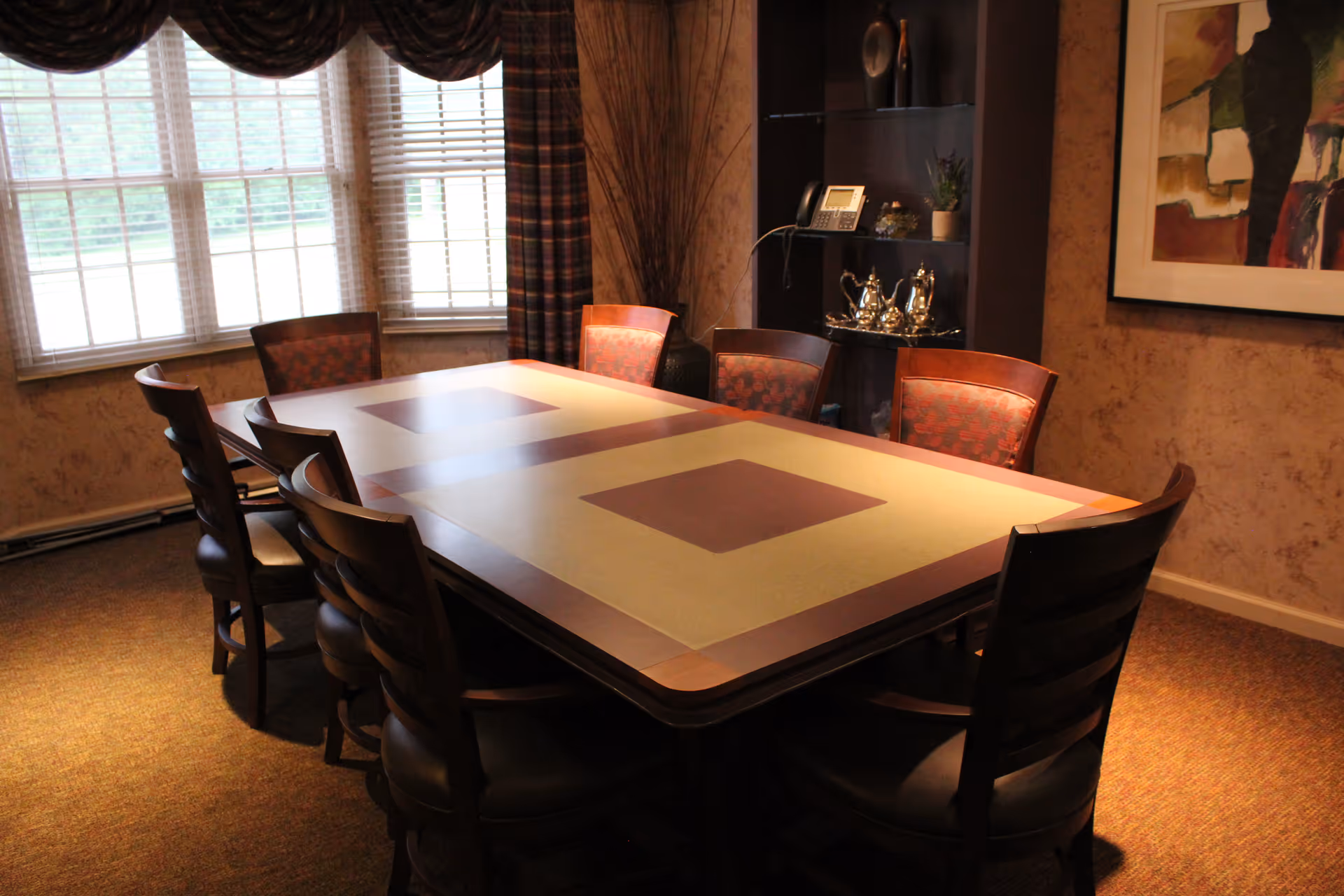 A dining room with a large rectangular table surrounded by eight wooden chairs with cushioned seats. The room has a window with blinds and curtains, a wall-mounted shelf with decorative items and a phone, and a framed abstract painting on the wall.