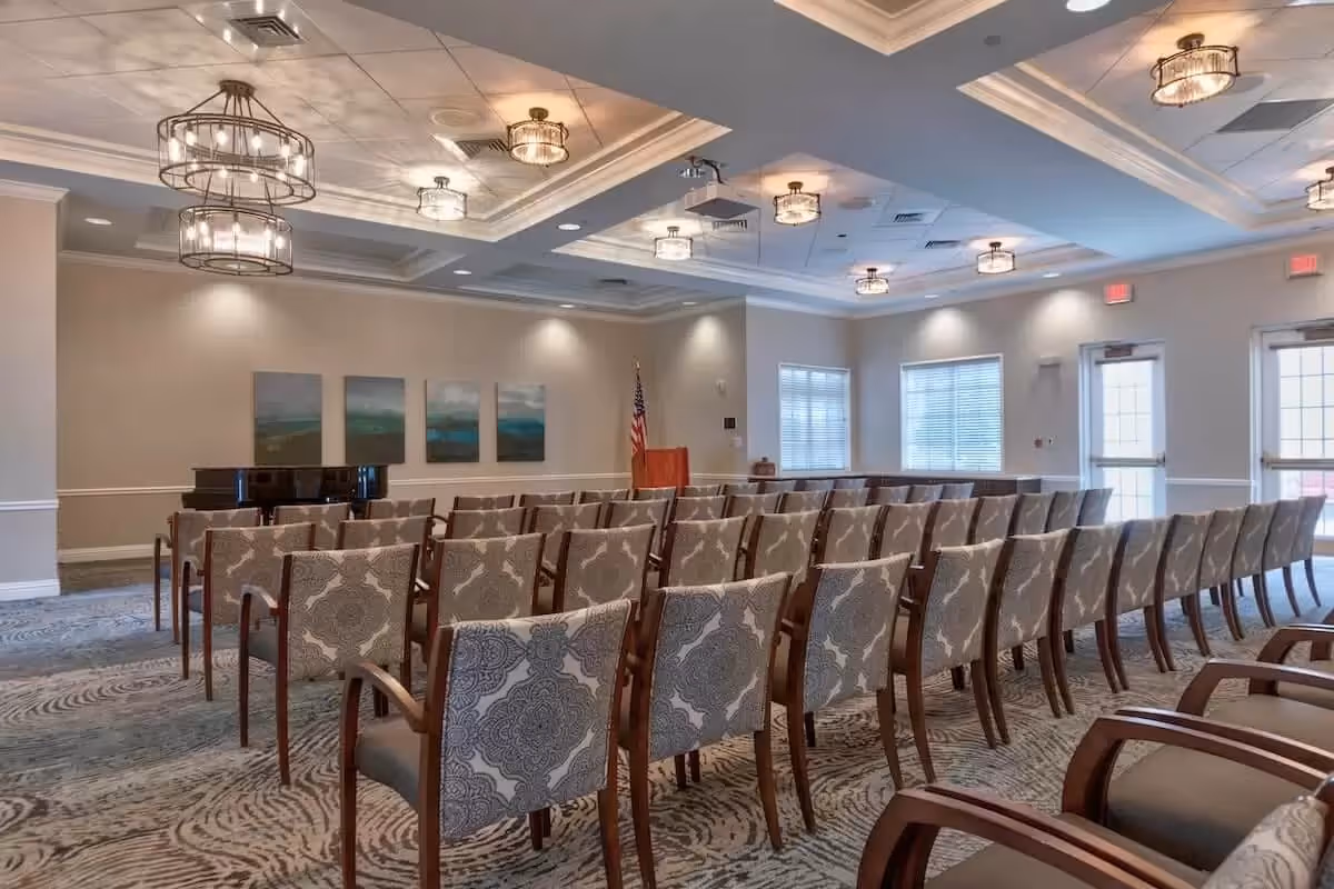 A well-lit meeting or event room with rows of patterned chairs facing a podium and a grand piano. The room has a decorative ceiling with multiple light fixtures, neutral-colored walls, and windows letting in natural light.