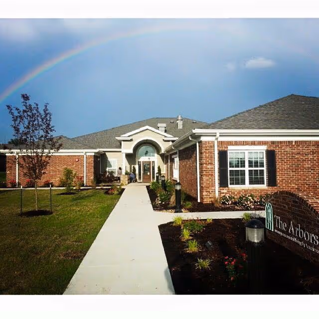 Exterior view of a single-story brick building with a gray shingled roof and white trim, featuring a central entrance with an arched window above the door. A concrete walkway leads up to the entrance, bordered by landscaped grass and small plants. A rainbow is visible in the sky above the building. A sign near the walkway reads 'The Arbors'.