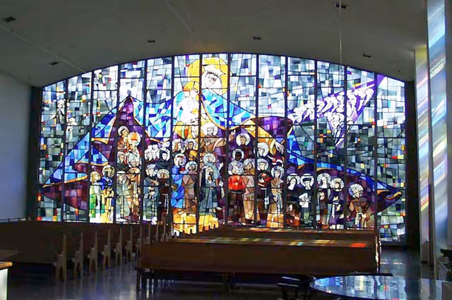 Interior chapel with rows of pews facing a large colorful stained-glass window depicting religious figures.