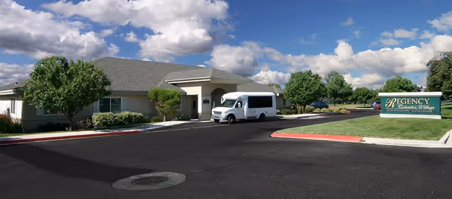 Exterior view of a single-story senior living facility building with a covered entrance and a white shuttle van parked in front. There are green trees and shrubs around the building, a clear blue sky with scattered clouds, and a sign on the right side that reads 'Regency at Columbia Village Assisted Living'.