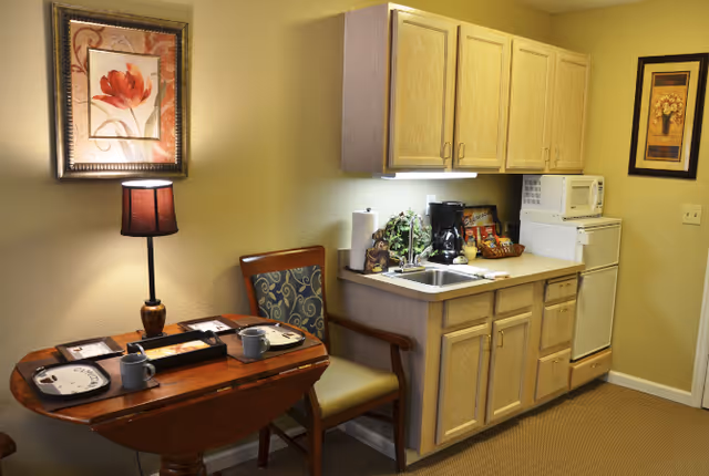 Small kitchenette and dining area with a table set for two, lamp, chair, sink, coffee maker, microwave, and cabinets.