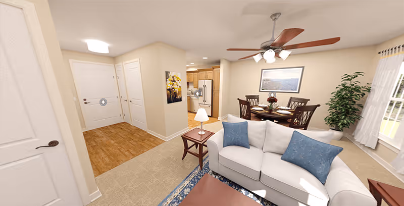 Interior view of a senior living community apartment showing a living room with a white sofa and blue cushions, a wooden coffee table, a side table with a lamp, a dining area with a round table and four chairs, a ceiling fan with lights, a large window with curtains, a potted plant, and a kitchen area with wooden cabinets and appliances in the background.