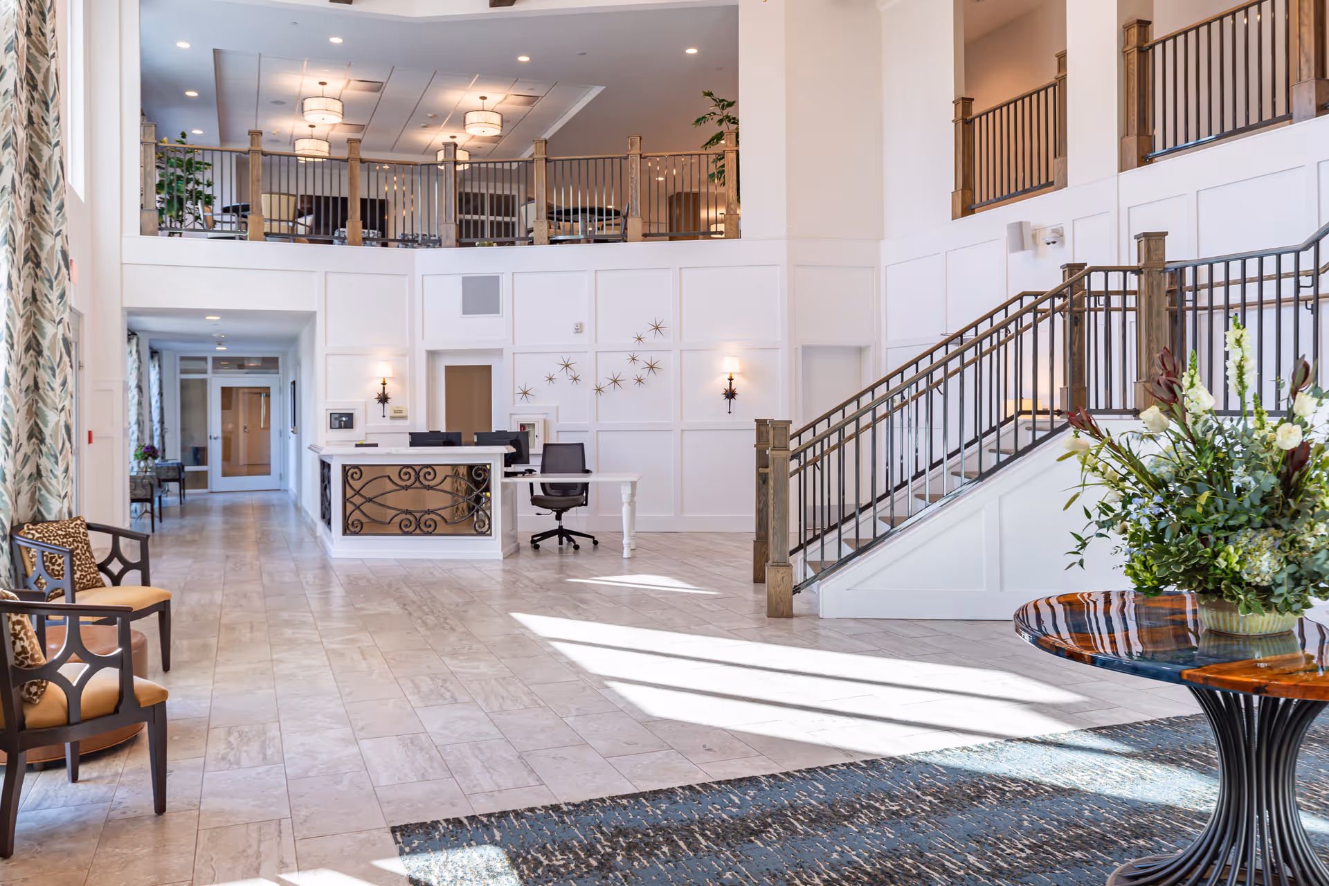 Bright and spacious senior living facility lobby with a reception desk, seating area with chairs, a staircase with wooden railings, and a round table with a large floral arrangement. The space features high ceilings, large windows with patterned curtains, and a second-floor balcony overlooking the lobby.