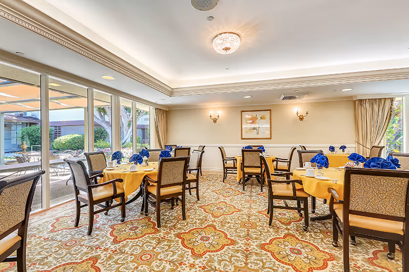A bright dining room with several round tables covered with yellow tablecloths and set with blue folded napkins, cups, and saucers. The room has large windows on one side showing an outdoor garden area, patterned carpet, beige walls with framed artwork, and elegant ceiling lighting.