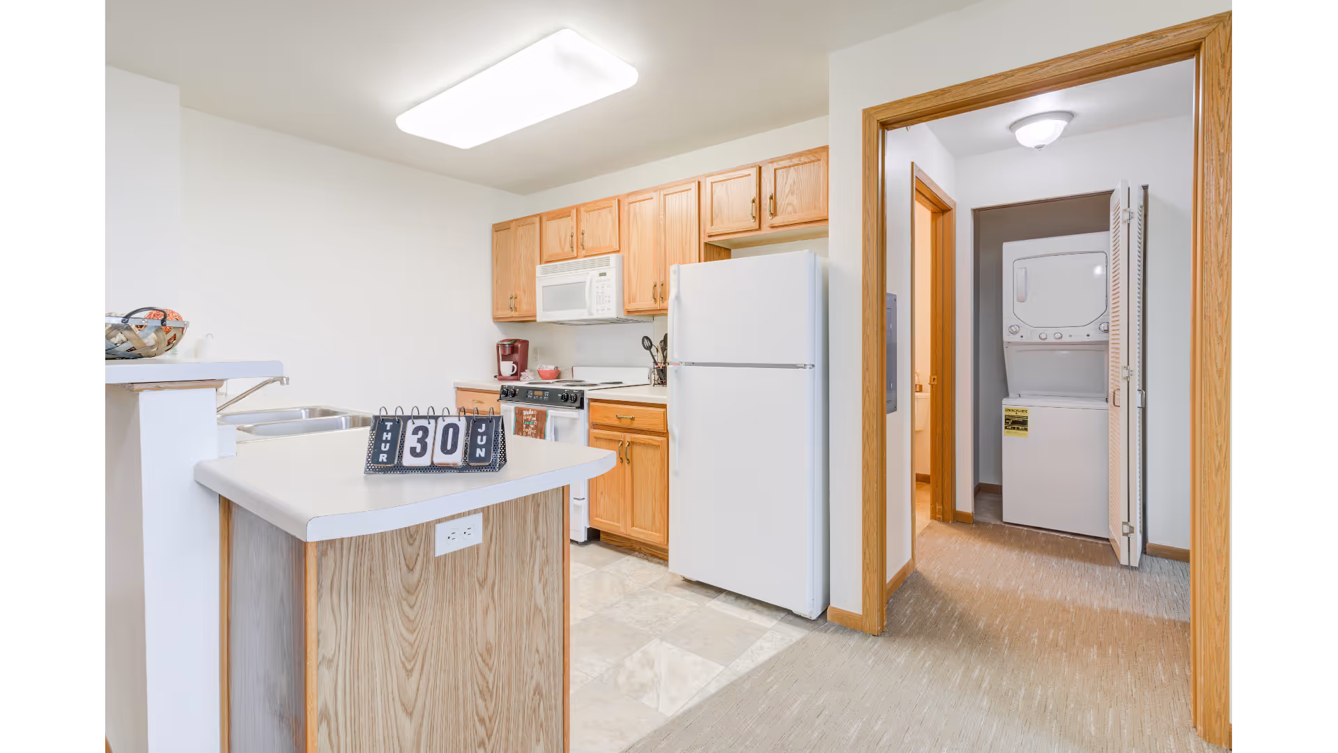 A bright kitchen area with wooden cabinets, a white refrigerator, a microwave above a stove, and a coffee maker on the counter. A small island with a calendar showing Thursday, June 30 is in the foreground. To the right, there is an open doorway leading to a laundry area with a stacked washer and dryer unit.