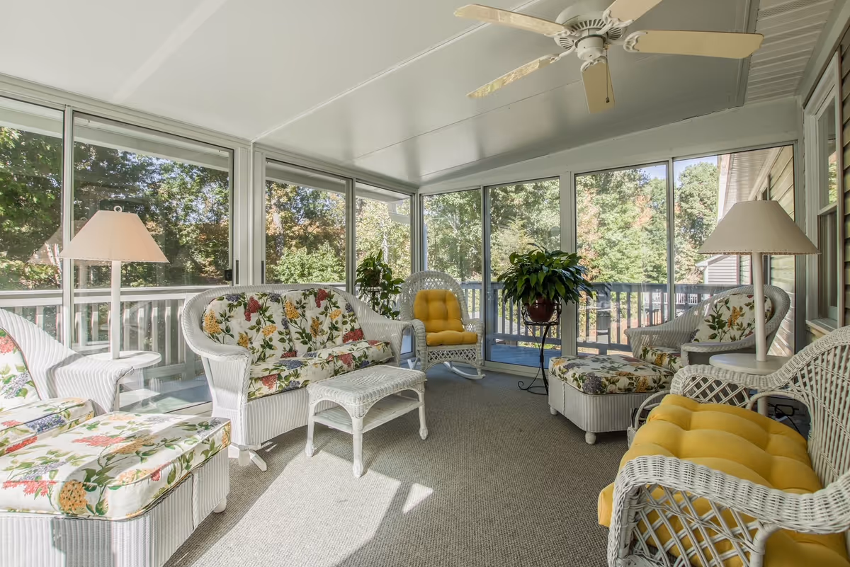 Bright screened-in sunroom with white wicker seating, floral cushions and yellow pads, a potted plant, and large windows showing trees.