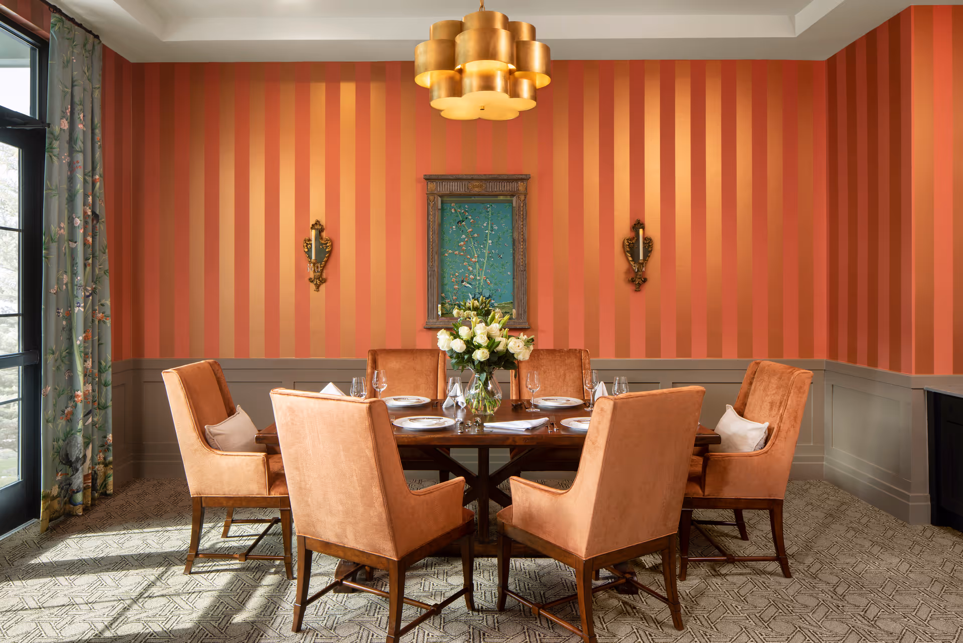 A dining room with a round wooden table set for six people with plates, glasses, and napkins. Six upholstered chairs in a warm orange color surround the table. The walls have vertical orange and red stripes with a gray wainscoting. A floral painting is centered on the wall, flanked by two ornate wall sconces. A gold chandelier hangs above the table, and a window with floral curtains allows natural light into the room.