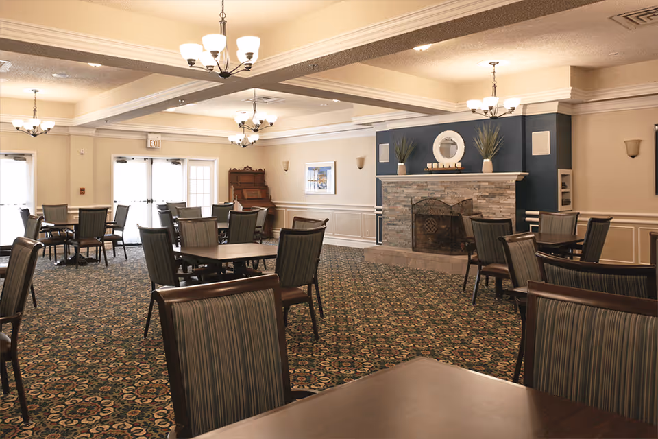 A spacious dining room with multiple tables and chairs arranged on a patterned carpet. The room features a stone fireplace with decorative plants and a round mirror above it. There are chandeliers hanging from the ceiling and large windows letting in natural light.