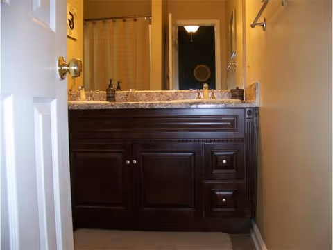 View of a bathroom vanity with a dark wood cabinet, granite countertop, and a sink with a faucet. A mirror is mounted above the sink, reflecting a hallway and a light fixture. The bathroom has beige walls and a towel rack on the right side.