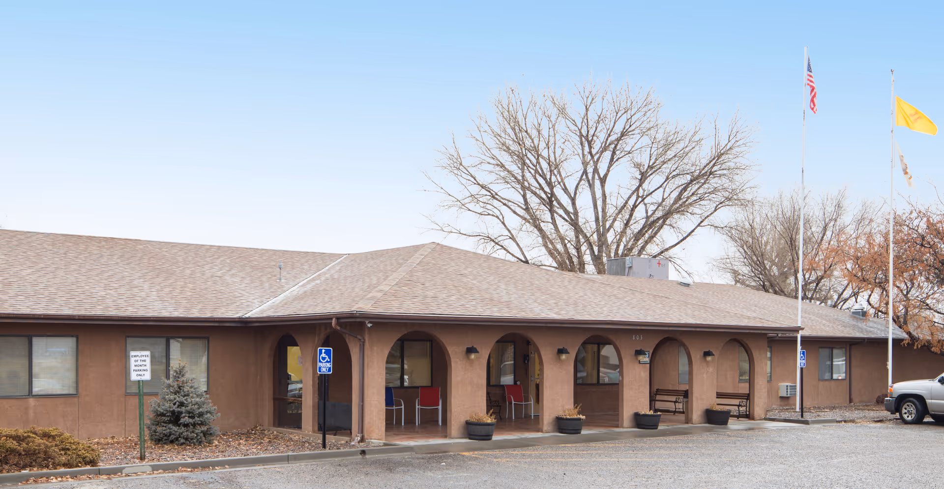 Single-story building with a brown exterior and arched entryways. There are several chairs and benches under the covered walkway. Two flagpoles with flags are visible on the right side, and a few leafless trees are in the background. A handicapped parking sign and a sign indicating employee parking only are visible in front of the building.