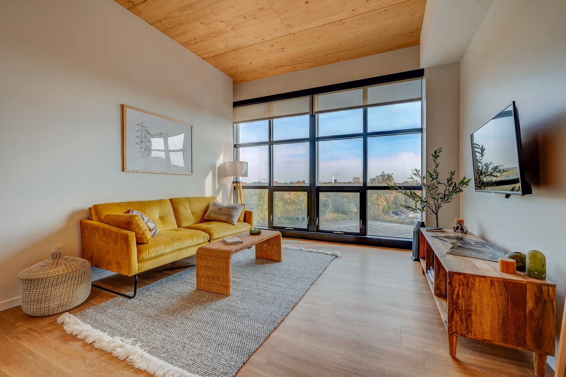 A modern living room with a large window offering a scenic view outside. The room features a mustard yellow sofa with cushions, a woven coffee table on a textured gray rug, a floor lamp, a wooden media console with decorative items, and a wall-mounted flat-screen TV. The ceiling is wooden, and the floor is light wood.