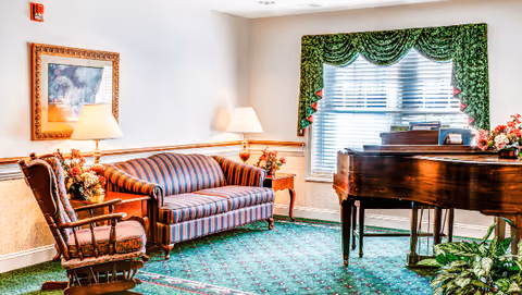 A cozy living room area with a striped sofa, a wooden rocking chair, two side tables with lamps and floral arrangements, a framed painting on the wall, a large window with green valance curtains, and a wooden grand piano with a floral arrangement on top.