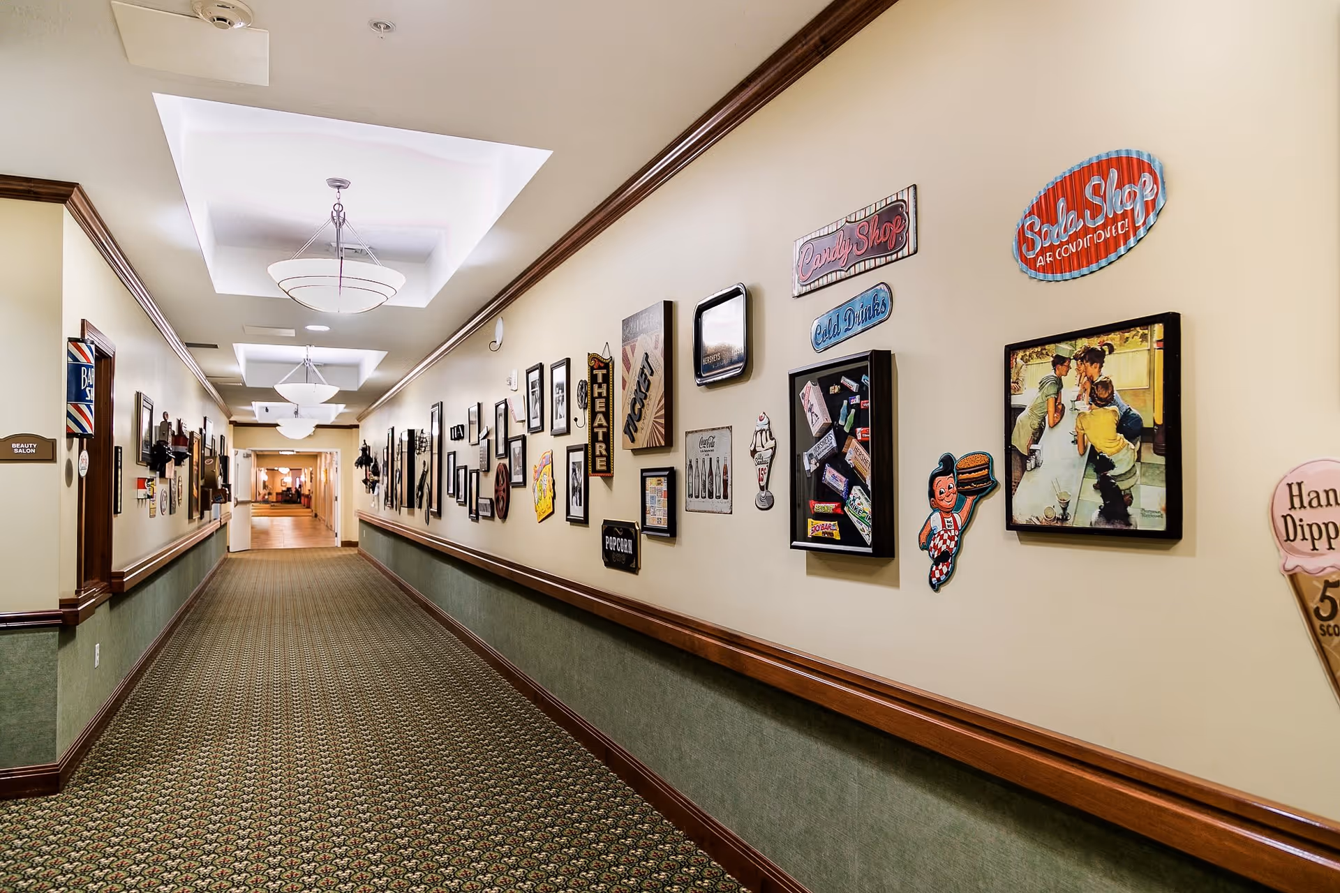 A long, carpeted hallway in a senior living facility with framed vintage-style signs and pictures decorating the right wall. The left wall has a door labeled 'Beauty Salon' and a barber pole sign. The ceiling has recessed lighting fixtures with decorative covers.
